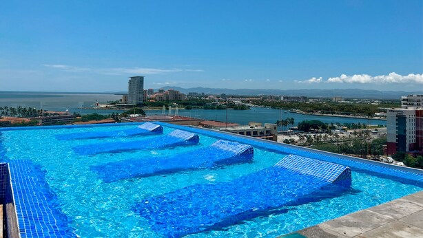 Uma piscina infinita no terraço, com água azul cristalina, rodeada por edifícios da cidade e vista para a costa.