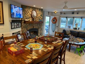 A dining area with a large wooden table set for board games, a stone fireplace, and a television mounted on the wall.