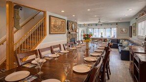 Een eetkamer met een lange tafel gedekt voor een maaltijd, een trap aan de linkerkant, en een zithoek met een bank en een open haard.