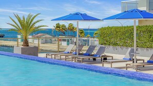 Un espace piscine doté de chaises longues et de parasols, avec vue sur une plage et l’océan.