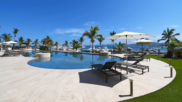A pool area with lounge chairs and umbrellas, surrounded by palm trees and a view of the sea.