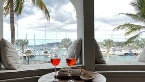 A view from a window with two glasses of red drink on a small table, overlooking a marina with boats and palm trees.