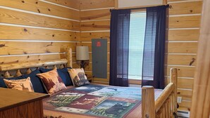 Ein Zimmer in einer Holzhütte mit einem Bett, einem Nachttisch, einer Lampe und einem Fenster mit Vorhängen.