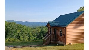 Eine Blockhütte mit Metalldeckung, einer Holzterrasse und Blick auf einen bewaldeten Gebirgszug.