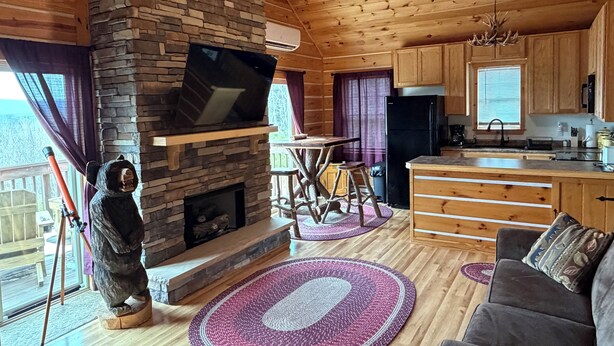 Ein gemütliches Blockhaushaus-Interieur mit Kamin aus Stein, einem an der Wand befestigten Fernseher, Holzdielen, einer Essecke und einer Küche.