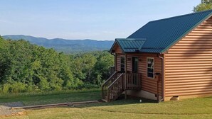 A log cabin with a metal roof, a wooden deck, and a view of a forested mountain range.