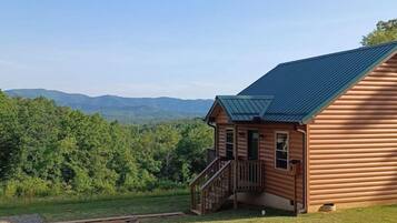Une cabane en rondins avec un toit en métal, une terrasse en bois et une vue sur une chaîne de montagnes boisées.