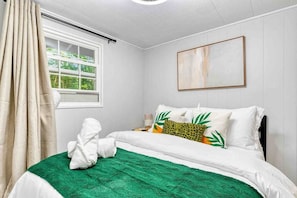 Cama bem arrumada com roupa de cama branca e um cobertor verde, uma janela com cortinas brancas e um quadro abstrato na parede.