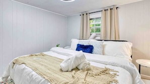 Una cama bien tendida con edredón blanco, una manta de color beige y una toalla blanca enrollada. Hay una mesita de noche con una lámpara y una ventana con cortinas color beige.