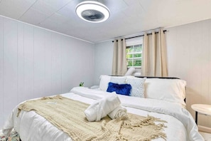 A neatly made bed with a white duvet, a beige throw blanket, and a rolled white towel. There is a small bedside table with a lamp, and a window with beige curtains.