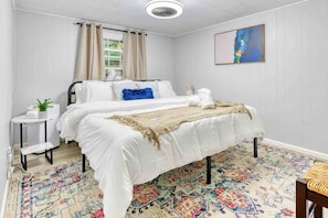 A bedroom with a bed, a small bedside table, a window with curtains, a colorful rug, and a painting on the wall.