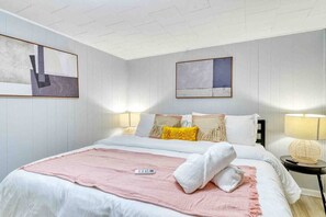 A hotel room with a large bed, a bedside table with a lamp, and two abstract paintings on the walls. - Red Roof Bungalow | Hot Tub Fire Pit Dog Friendly (Coolbaugh Township)