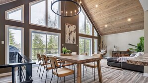 A spacious living area with a large wooden dining table, multiple chairs, a hanging light fixture, and a view through large windows.