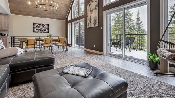 A spacious living area with a high ceiling, a dining set, and a balcony with a view of trees.