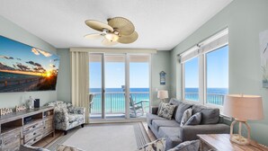 A spacious living room with a large window offering a view of the ocean, a ceiling fan, and a painting of a sunset. - 🌞Beachfront|Gulf Views|Pools|3BR Summerwind 804🌞 (Navarre Beach)