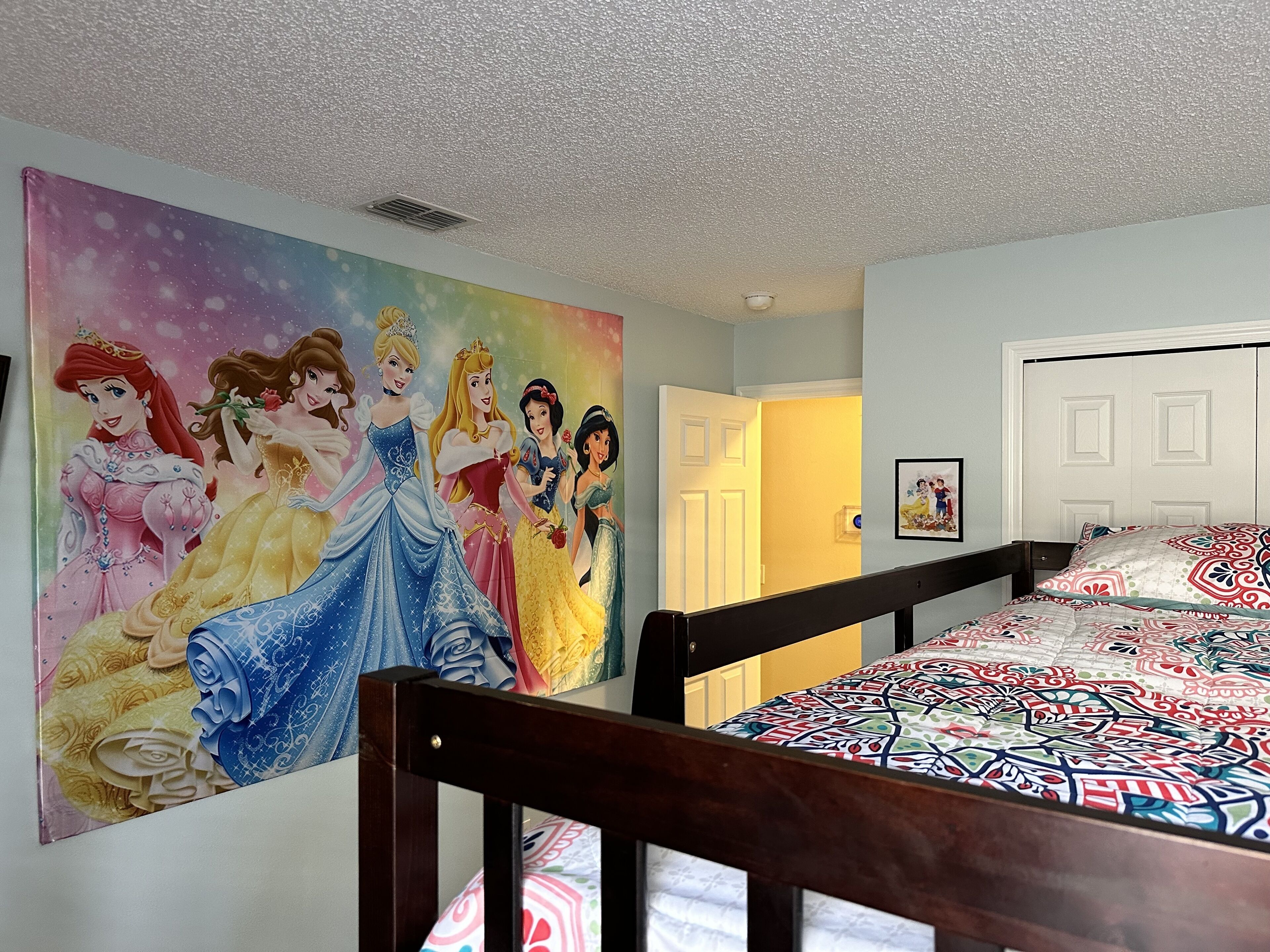A bedroom with a large Disney princess mural on the wall, a bed with a patterned bedspread, and a door leading to another room.