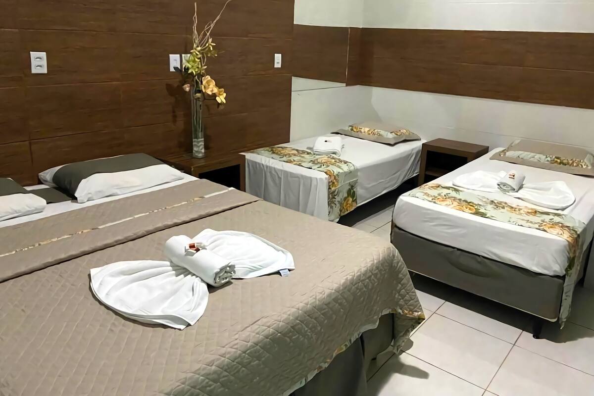A hotel room with three beds, each with a white bedspread and pillows. There is a vase with flowers on the nightstand between the beds.