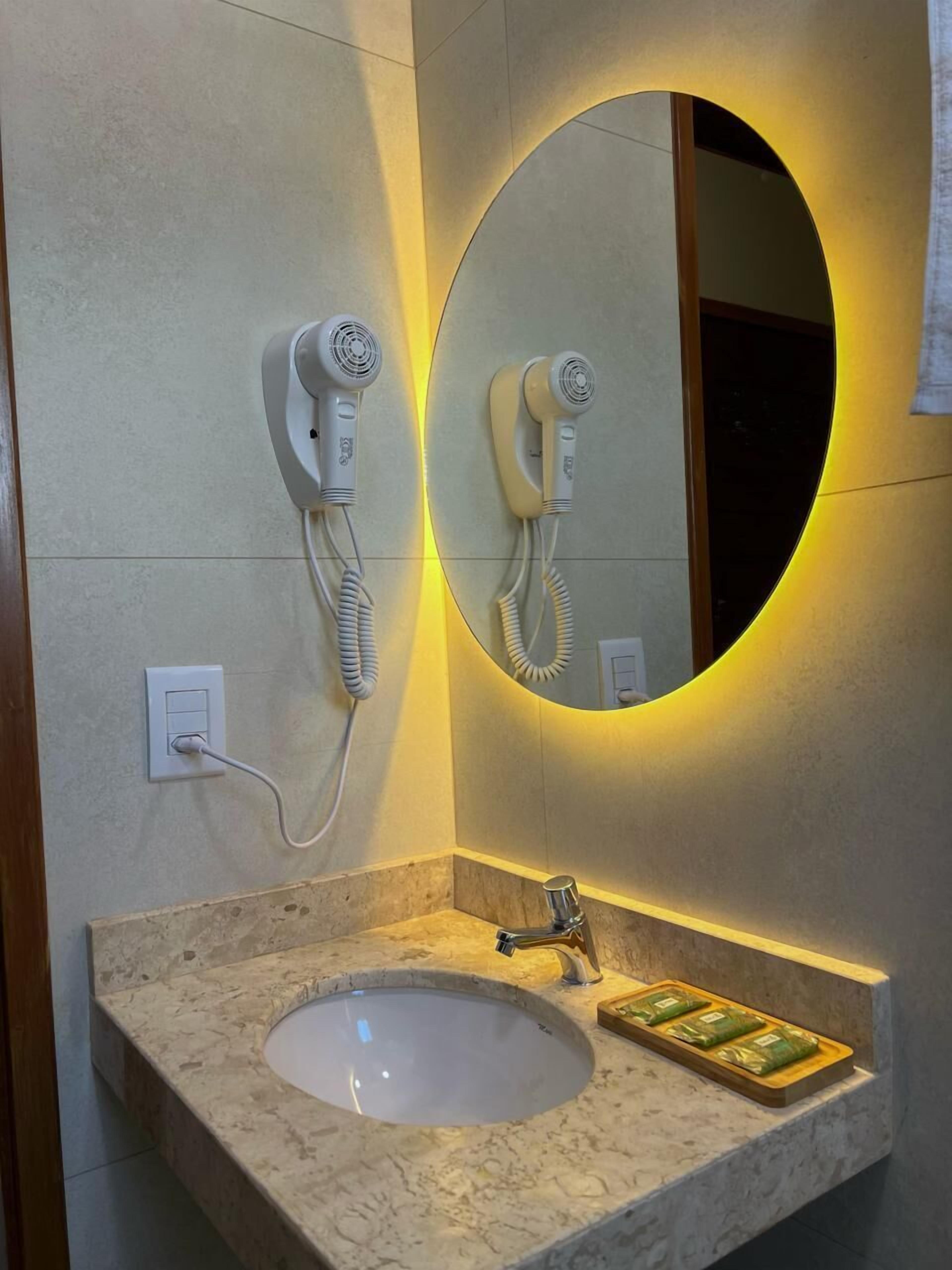 Un baño con lavamanos de mármol, un espejo con luz circular y dos secadores de pelo fijados a la pared.