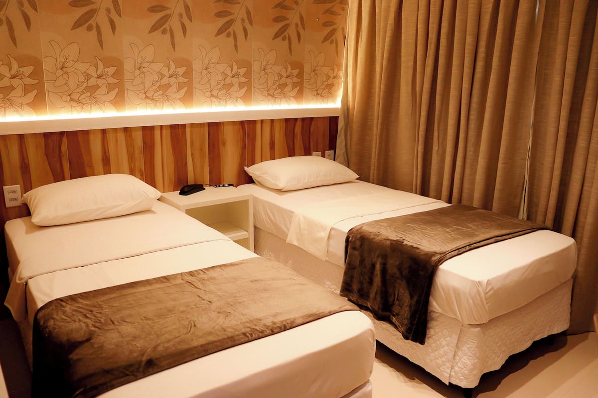 A hotel room with two single beds, a nightstand, and a wall with floral wallpaper.