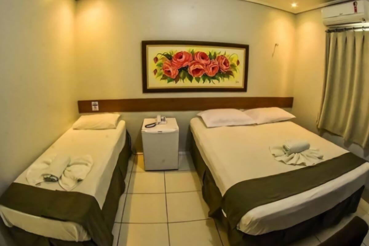 A hotel room with two beds, a small bedside table with a phone, a painting of flowers on the wall, and an air conditioning unit.