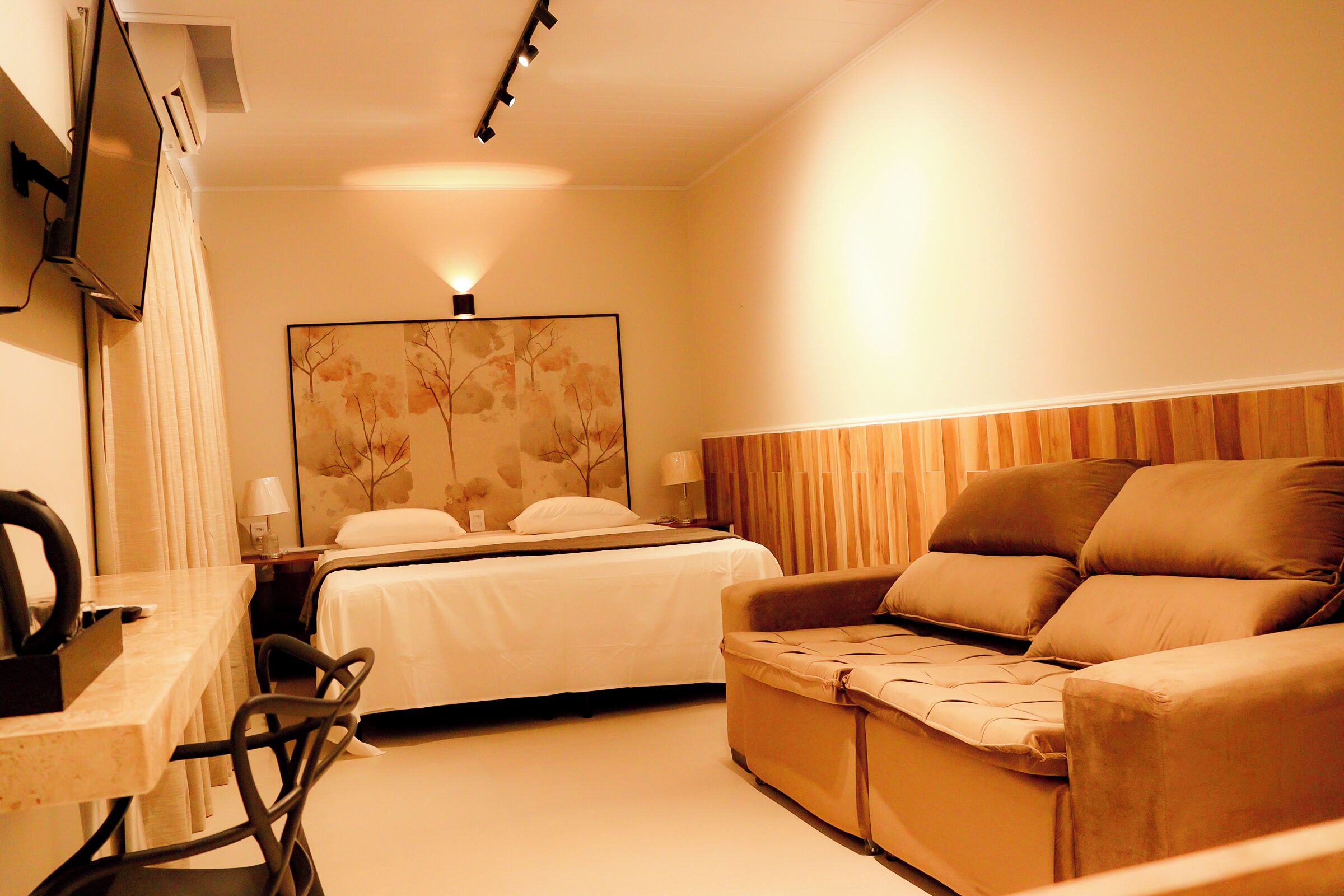 A hotel room with a bed, a sofa, a desk with a chair, a TV mounted on the wall, and a painting on the wall.