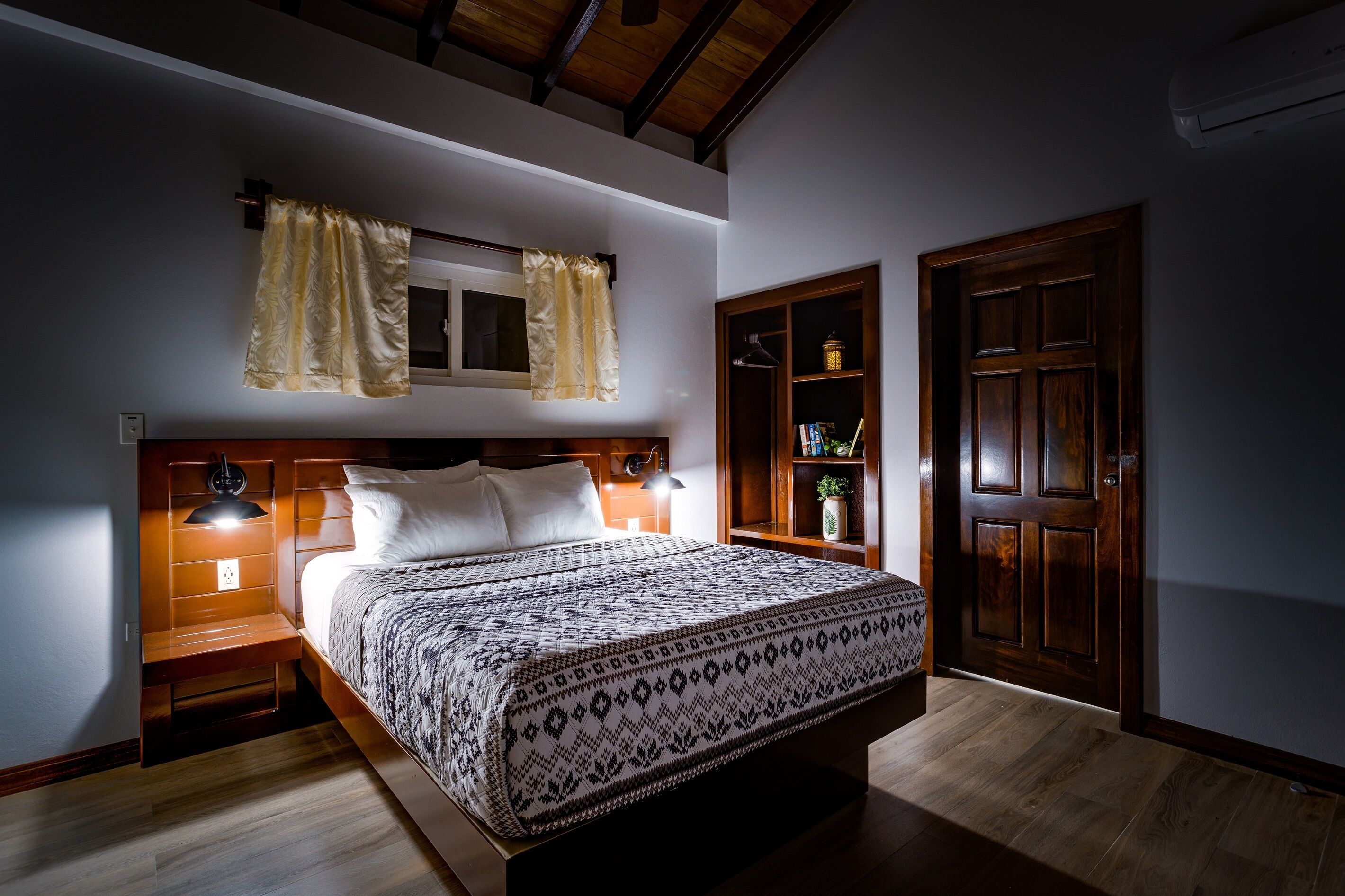 A bedroom with a wooden bed, a nightstand, a bedside lamp, a wardrobe, and a window with curtains.