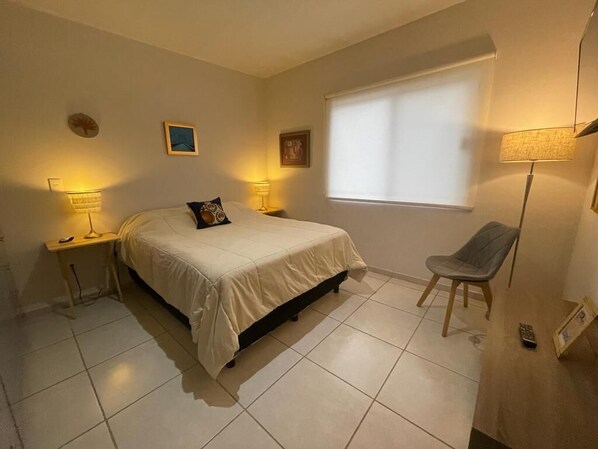 A bedroom with a bed, two bedside lamps, a chair, a window, and a wall clock. - Condo en Residencial con Alberca (Durango)