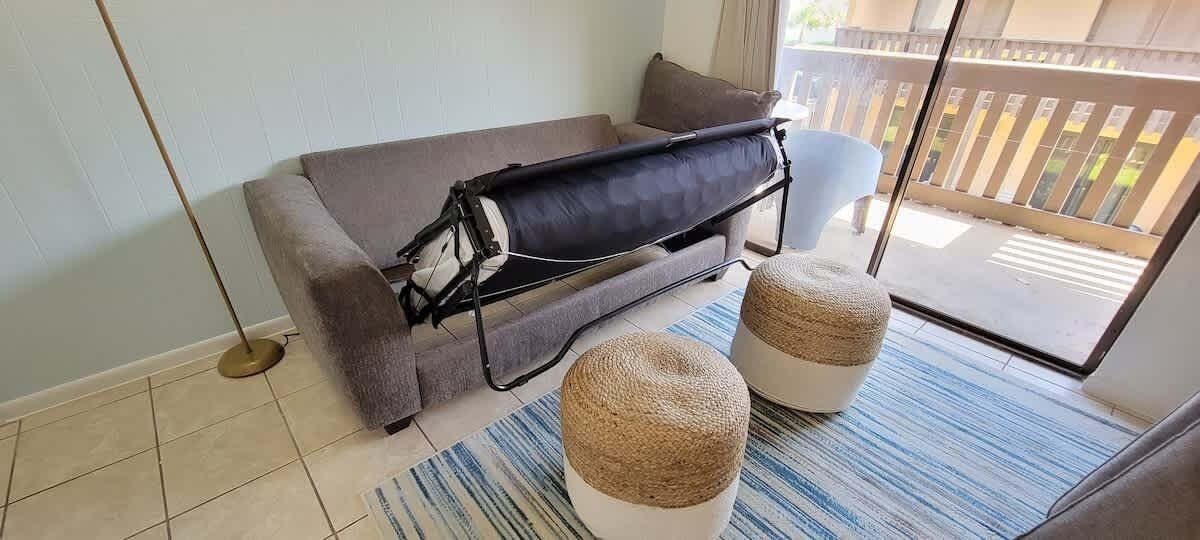 Une pièce avec un canapé gris, un matelas noir sur roulettes, deux poufs ronds, un tapis à rayures et une lampe sur pied.