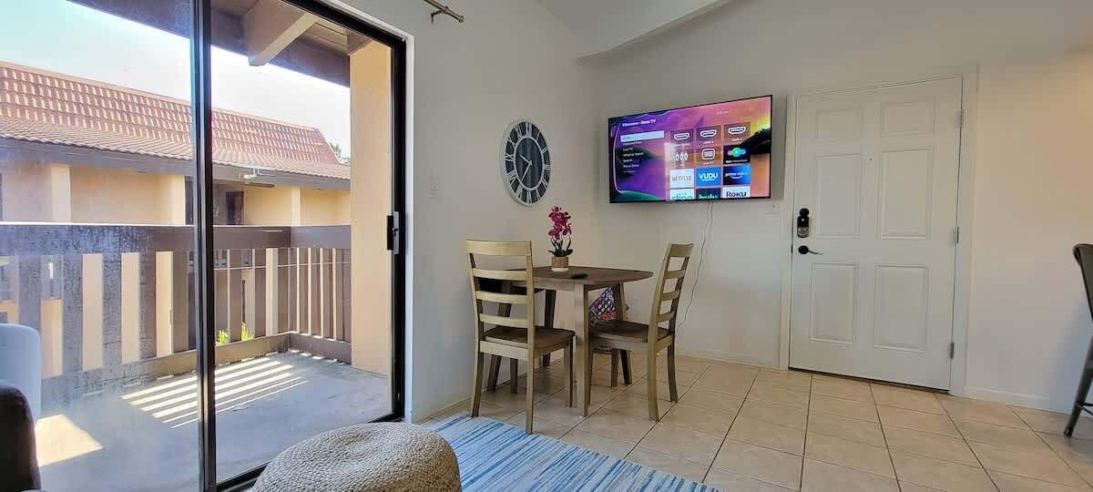 Ein Balkon mit einer Schiebetür, ein Essbereich mit Tisch und Stühlen, ein an der Wand befestigtes Fernsehgerät und eine Tür mit Griff.