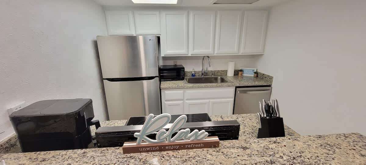 A kitchen with white cabinets, a stainless steel refrigerator, a granite countertop, and a sign that reads 'Relax.'