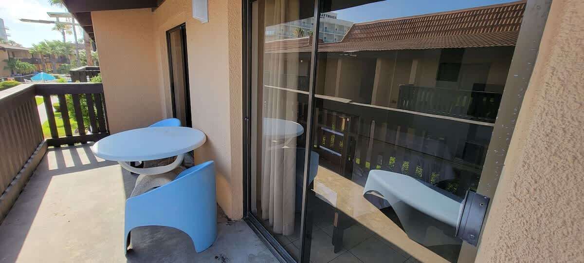Un balcon avec une table blanche et des chaises bleues, une porte-fenêtre coulissante et une vue sur un immeuble avec un toit en tuiles.