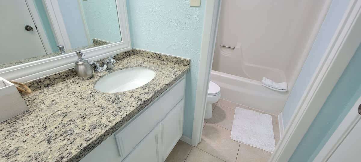 Un baño con lavamanos de encimera de granito, un espejo, inodoro y zona de ducha.