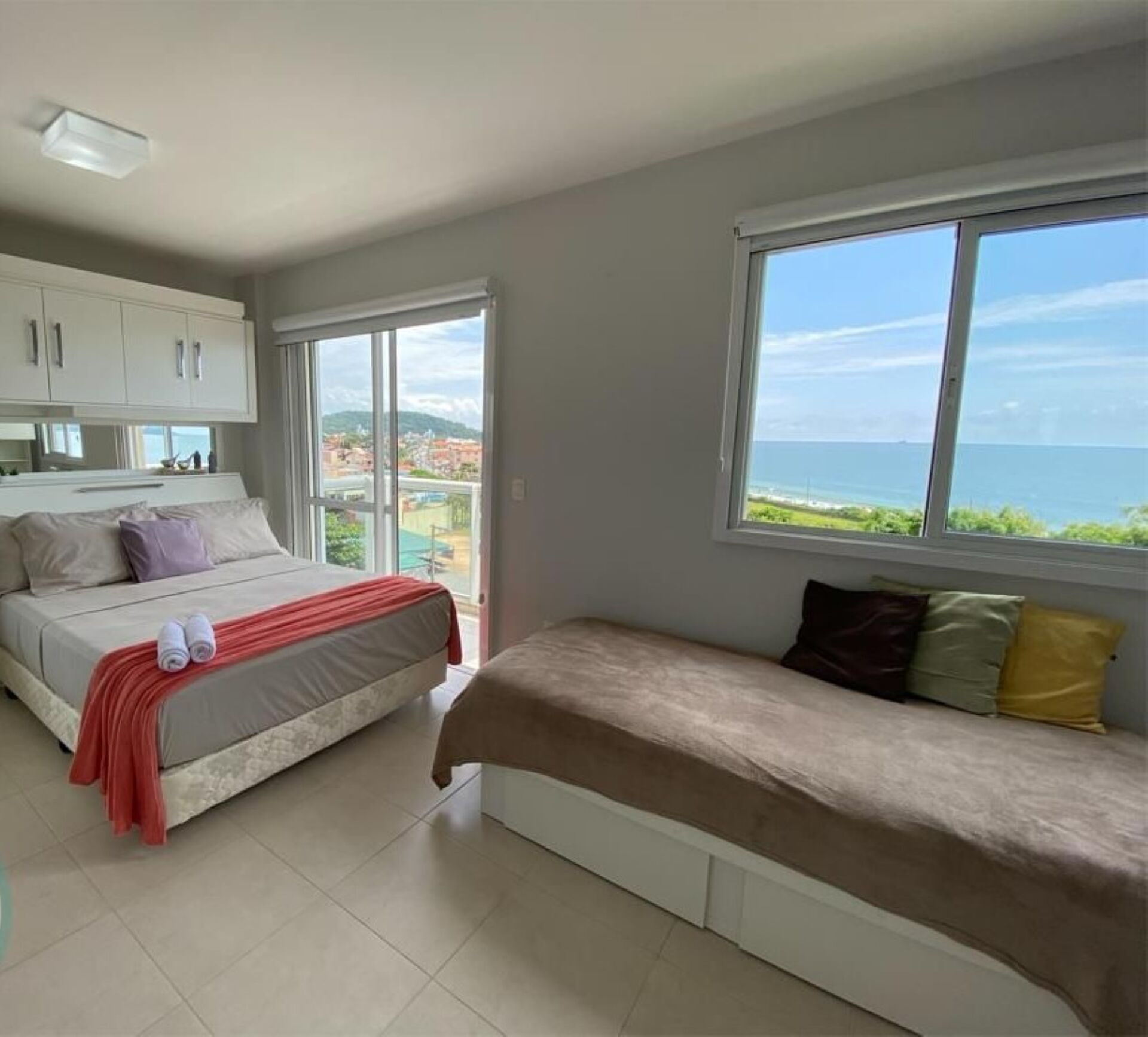 A bedroom with a bed, a sofa, a window with a view of the sea, and a kitchenette.