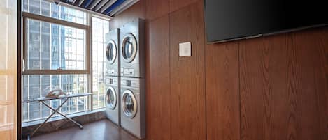 A laundry room with four washing machines, a large window, and a flat-screen TV mounted on the wall.