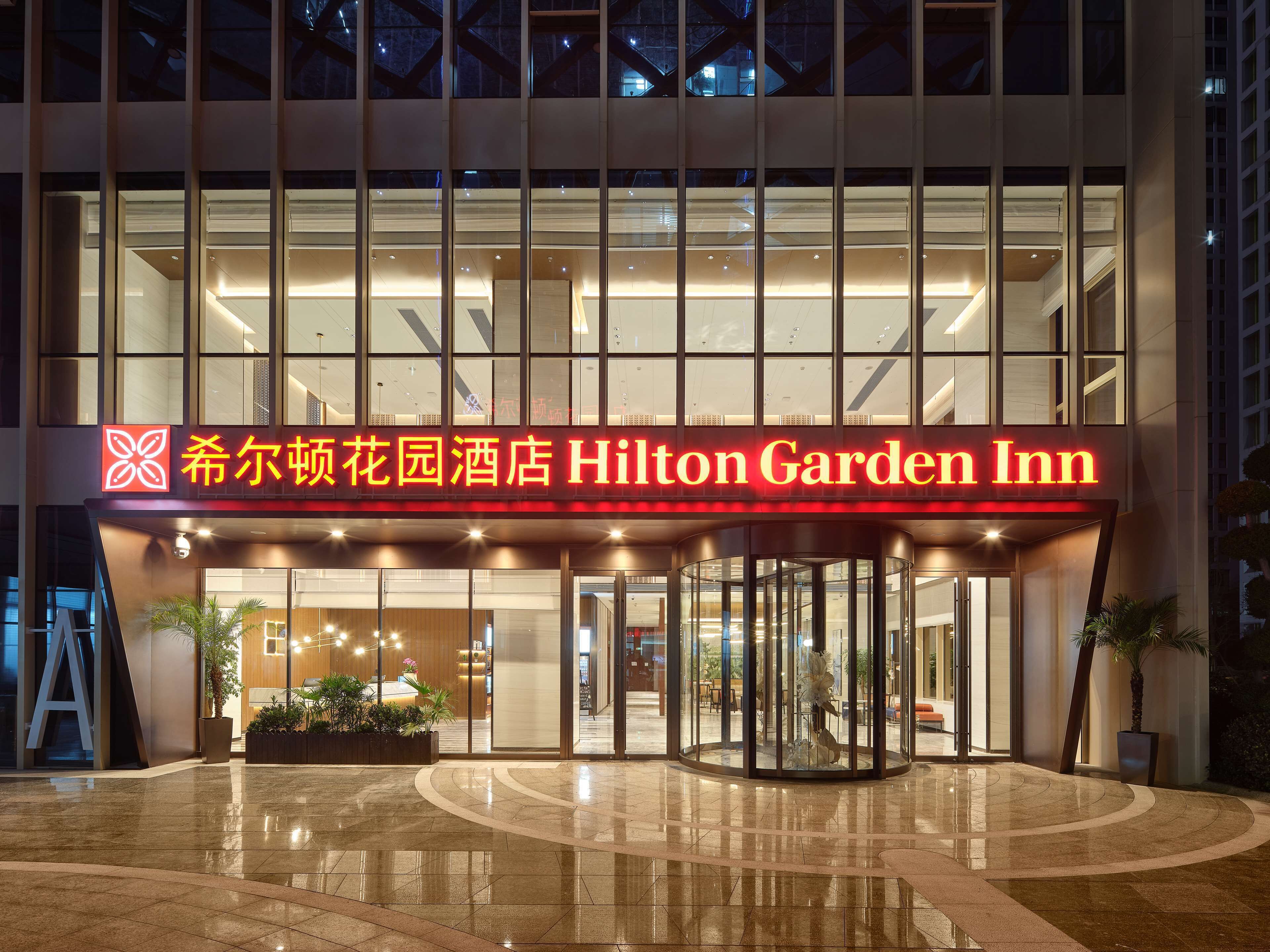 L’extérieur de l’hôtel Hilton Garden Inn se distingue par sa façade moderne en verre, son entrée imposante avec des portes tournantes et un panneau indicateur bien éclairé.
