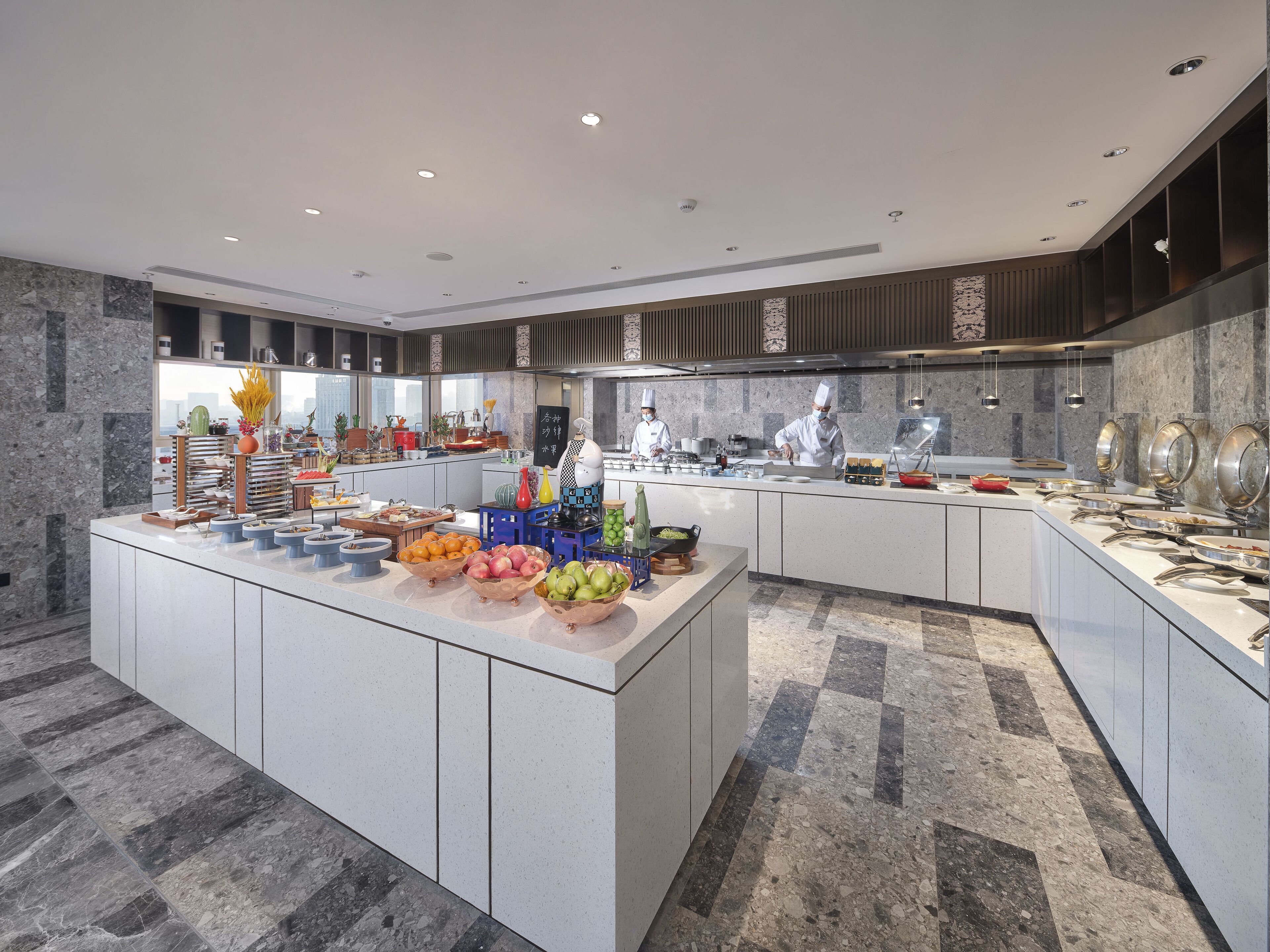 Una cocina de hotel moderna con una isla central para bufé, pisos de mármol y vistas de la ciudad a través de amplias ventanas.
