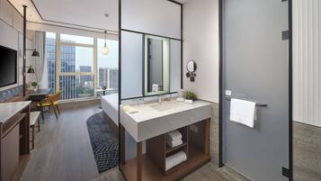 Ein modernes Hotelzimmer mit einem großen Bett, einem Badezimmer mit großem Spiegel und Waschbecken, einer Essecke mit Tisch und Stühlen sowie Blick auf die Stadt durch große Fenster.