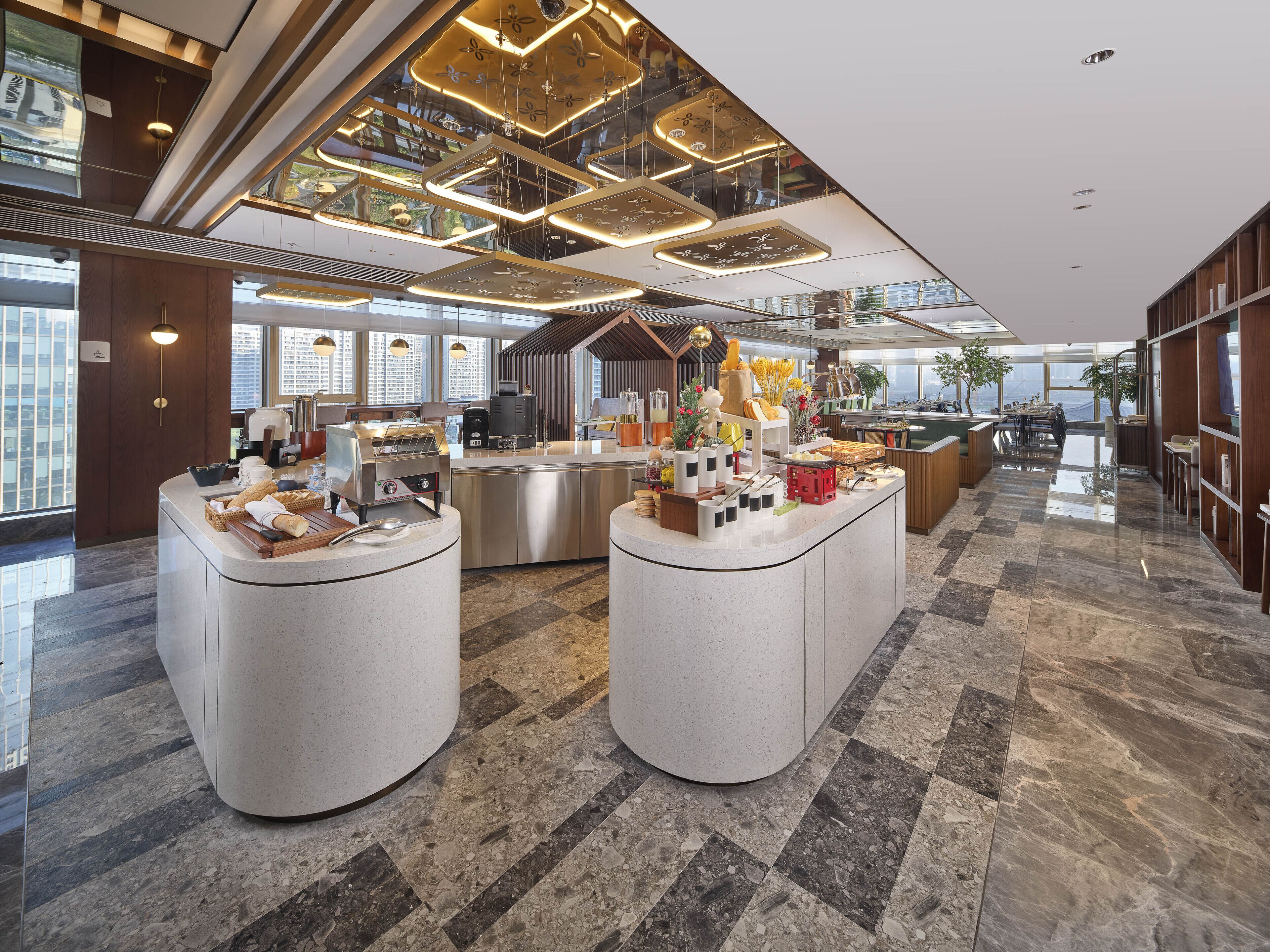 A modern hotel lobby with two central round counters, a coffee station, and a seating area with a view of the cityscape.