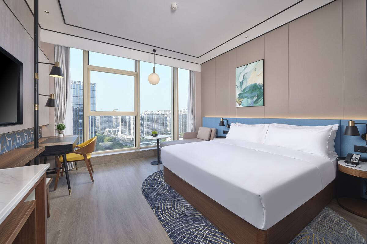 Ein modernes Hotelzimmer mit einem großen Bett, einem Schreibtisch, einem Sessel, einem kleinen Tisch mit Pflanze, einem Fernseher und Blick auf die Stadt durch große Fenster.