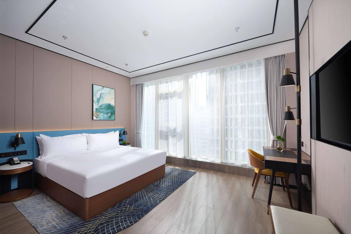 Ein modernes Hotelzimmer mit einem großen Bett, einem Schreibtisch, einem Sessel, einem Fernseher und einem Fenster mit Vorhängen.