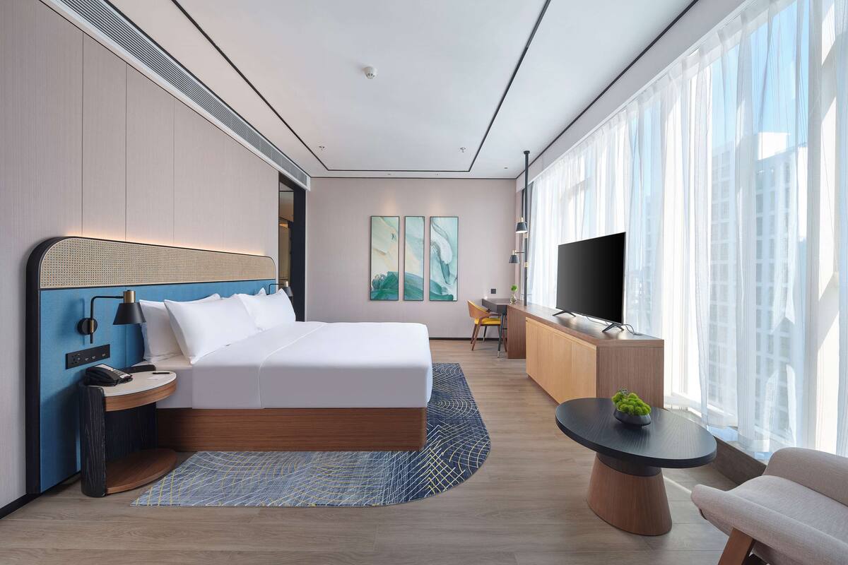Ein modernes Hotelzimmer mit einem großen Bett, einem Schreibtisch, einem Fernseher und Blick auf die Stadt durch große Fenster.