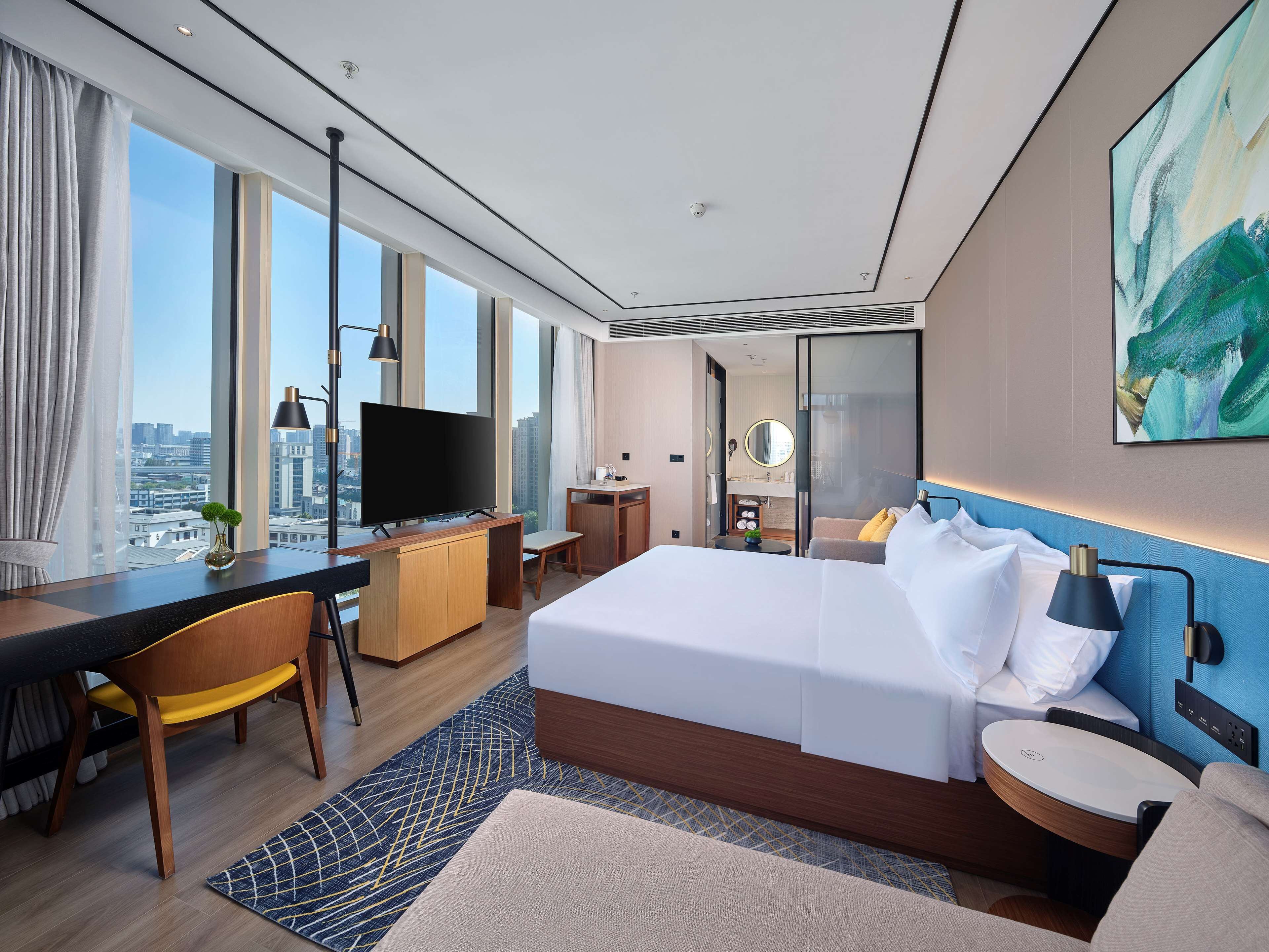 Une chambre d’hôtel moderne dotée d’un grand lit, d’un bureau avec un téléviseur à écran plat, d’une petite table et offrant une vue sur la ville grâce à de grandes fenêtres.