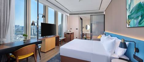 A modern hotel room with a large bed, a desk with a flat-screen TV, a small table, and a view of the city through large windows.