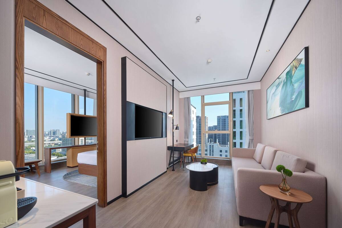 Ein modernes Hotelzimmer mit großem Fenster, einem Flachbildfernseher, einer Couch, einem kleinen Tisch und einem Sessel.