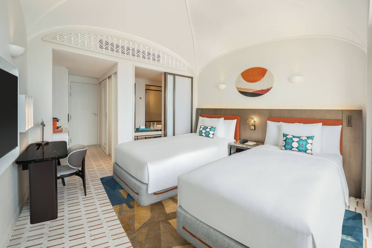 A hotel room with two beds, a desk, a chair, and a TV.