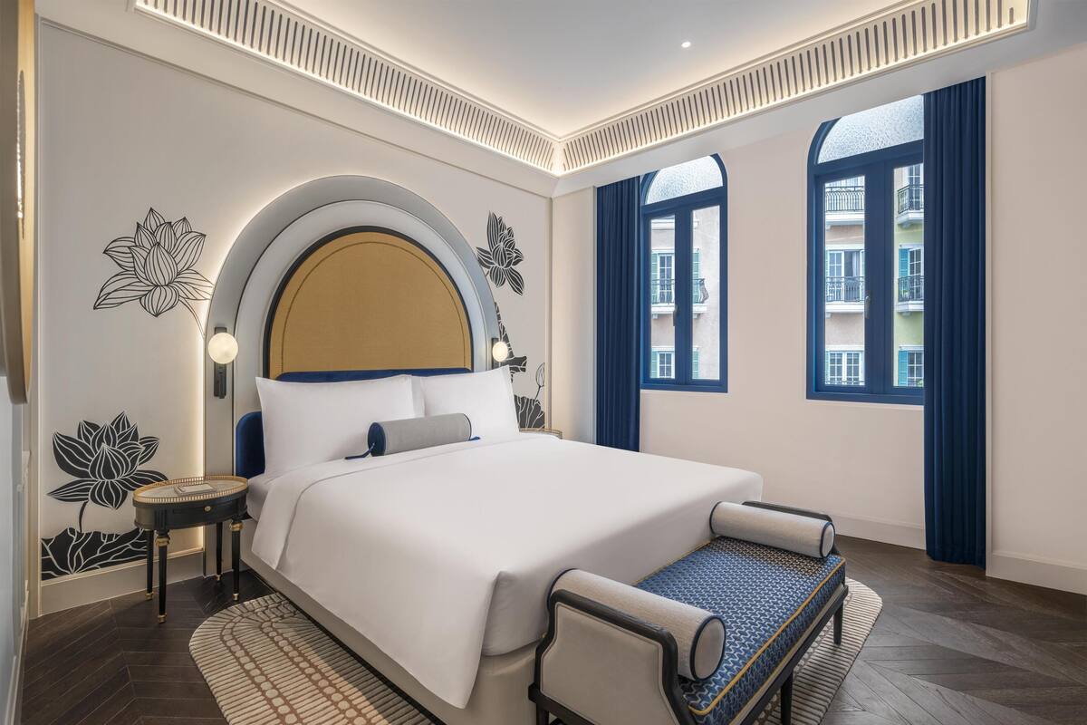 A modern hotel room with a large bed, a round side table, a small sofa, and a view of the city through large windows.