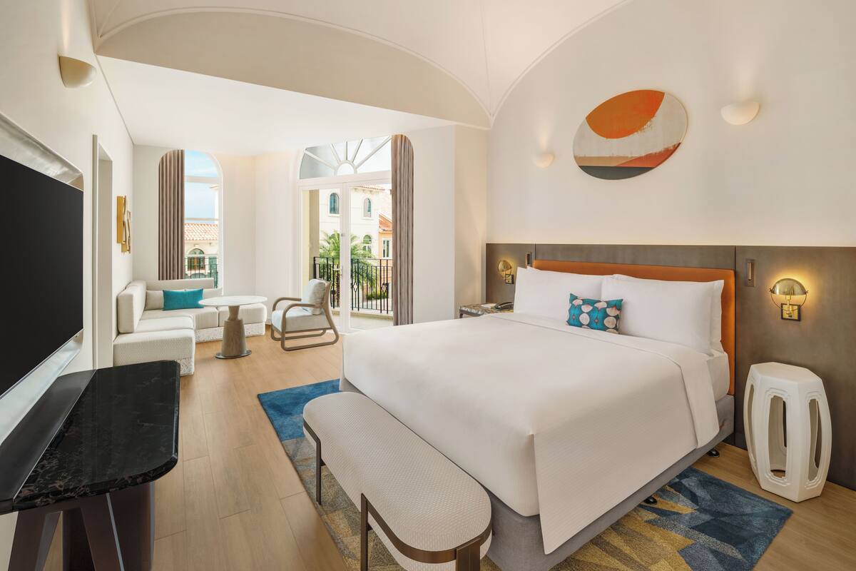 A hotel room with a bed, a desk, a chair, a bench, a TV, and a balcony with a view.