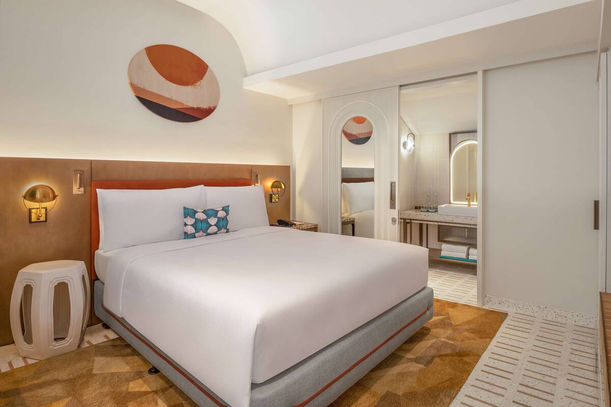 A hotel room with a large bed, a bedside table, a wall-mounted lamp, and a bathroom visible through an open door.