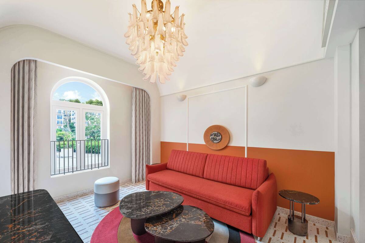 A modern living room with a red sofa, two black marble coffee tables, a chandelier, and a window with a view of greenery.