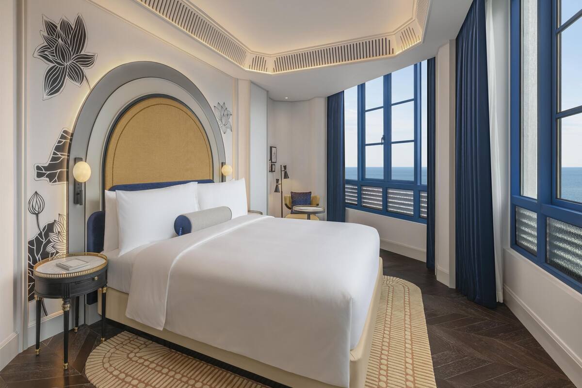 A modern hotel room with a large bed, a round side table, a small table, a decorative wall piece, and a view of the sea through large windows.